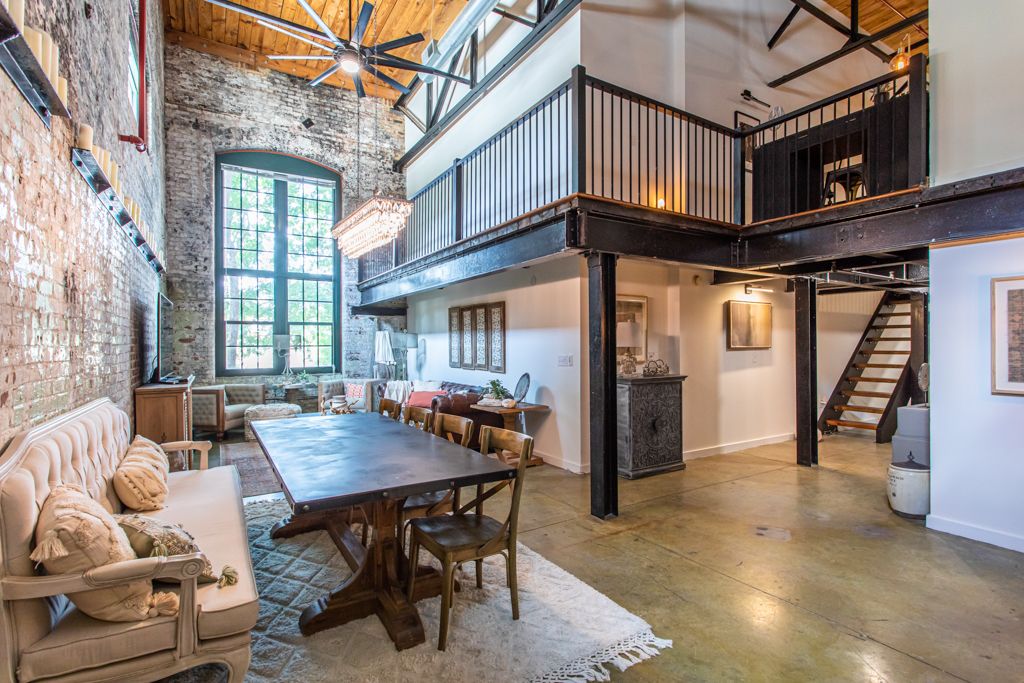 Double-height living and dining space with exposed brick, an arched factory window and the mezzanine above