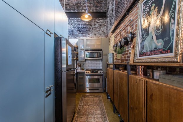 Galley kitchen with blue-grey cabinetry, stainless appliances and an exposed brick wall